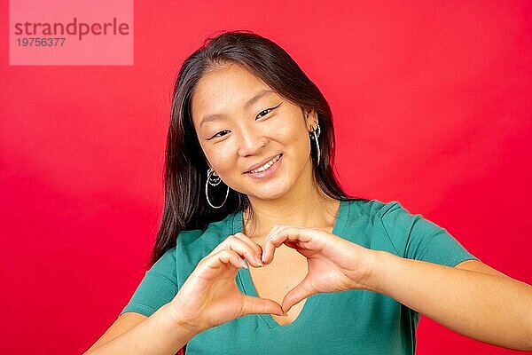 Studiofoto mit rotem Hintergrund einer chinesischen Frau  die sich die Hände reicht  um eine Herzform zu symbolisieren