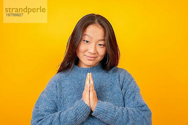 Asiatische Frau  die die Bitte Geste macht und in die Kamera schaut  auf einem gelben Hintergrund