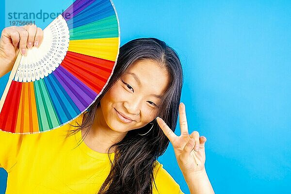 Studiofoto mit blauem Hintergrund von einer chinesischen Frau mit einem Regenbogen farbigen Fächer Geste Frieden