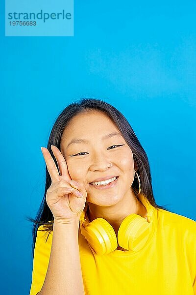 Vertikales Studiofoto mit blauem Hintergrund einer chinesischen Frau mit Kopfhörern  die ein Siegeszeichen gestikuliert