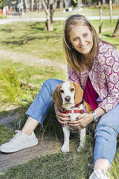 Glückliche blonde Frau im mittleren Erwachsenenalter  die für die Kamera lächelt und einen Beagle Hund umarmt  während sie auf grünem Gras im Park sitzt