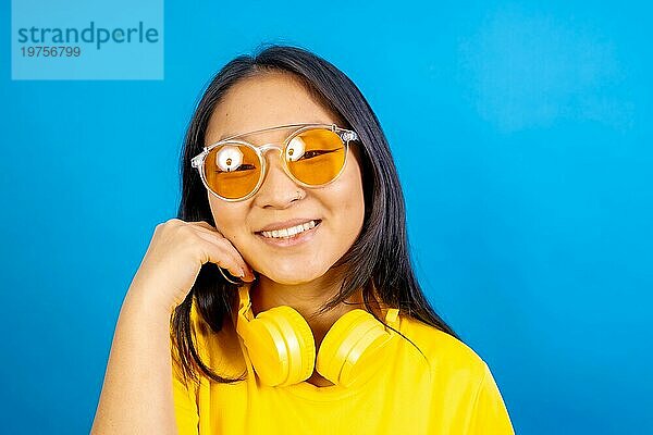 Studio Foto mit blauem Hintergrund einer schüchternen und niedlichen chinesischen Frau mit Sonnenbrille lächelnd in die Kamera