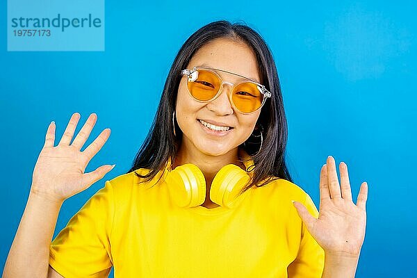 Studiofoto mit blauem Hintergrund einer glücklichen chinesischen Frau mit Kopfhörern im Nacken  die eine Sonnenbrille trägt und die Hände zum Gruß hebt