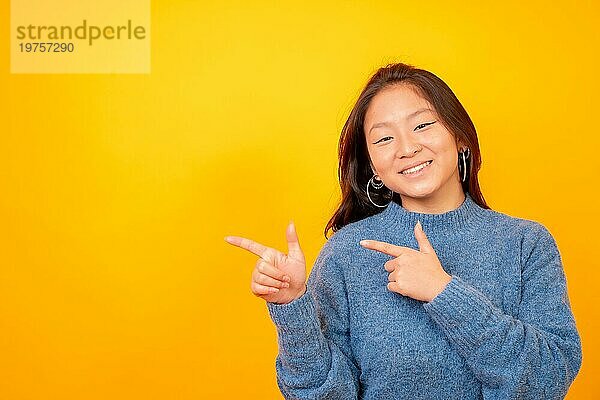 Studiofoto mit gelbem Hintergrund von einer glücklichen chinesischen Frau  die in einem leeren Raum zur Seite zeigt