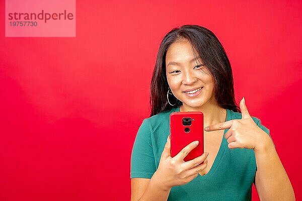 Studiofoto mit rotem Hintergrund einer chinesischen Frau  die auf ein Mobiltelefon zeigt