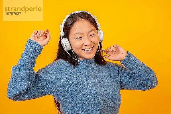 Asiatische Frau tanzt mit weißen Kopfhörern und schaut in die Kamera auf einem gelben Hintergrund