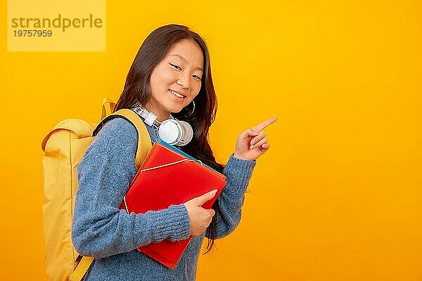 Studiofoto mit gelbem Hintergrund eines chinesischen Studenten mit Ordnern  Tasche und Kopfhörern  die auf einen schwarzen Raum zeigen