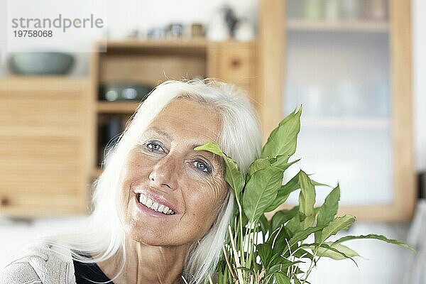 Ältere Frau mit langen Haaren schmiegt sich an Zimmerpflanze zu Hause
