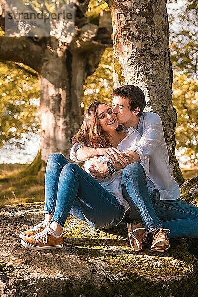Vertikales Foto eines jungen Mannes  der seine Freundin umarmt und küsst  während er im Wald sitzt