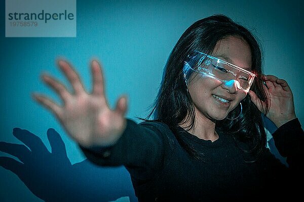Studiofoto mit blauem Hintergrund mit Neonlichtern einer chinesischen Frau  die mit einer Augmented Reality Brille gestikuliert und Spaß hat