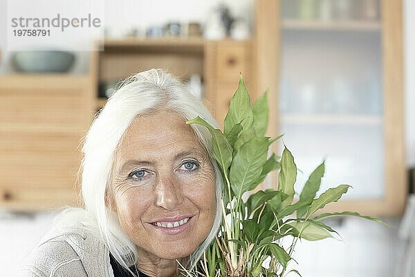 Ältere Frau mit langen Haaren schmiegt sich an Zimmerpflanze zu Hause