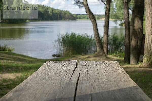 Alter  rissiger  verwitterter Picknicktisch aus Holz im Freien mit Blick auf einen ruhigen See oder Fluss mit schattigen Bäumen