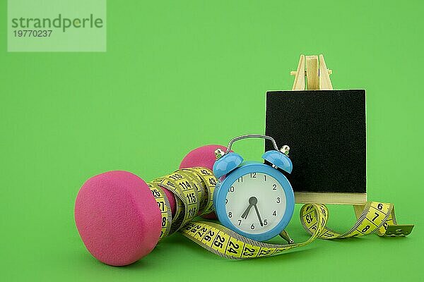Start gesunder Lebensstil Konzept. Stillleben mit rosa Hantel  Maßband  Kreidetafel und Wecker auf grünem Hintergrund