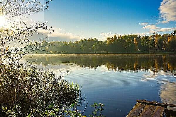 Sonnenuntergang über einem ruhigen ländlichen See mit Spiegelungen auf dem Wasser der umliegenden Bäume und Wälder mit einem rustikalen Holzsteg im Vordergrund und Nebel in einer malerischen Landschaft