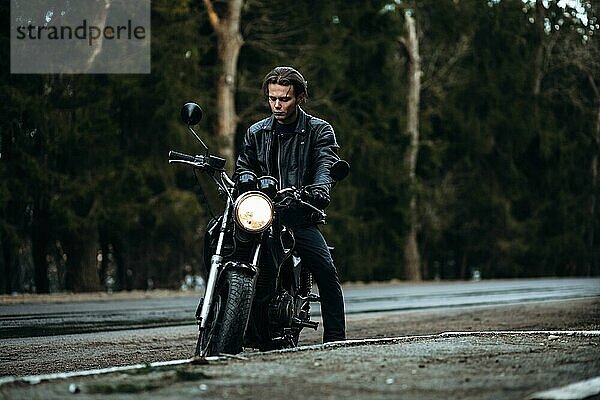 Stilvoller männlicher Motorradfahrer in einer Lederjacke mit einem klassischen Motorrad auf einer asphaltierten Waldstraße