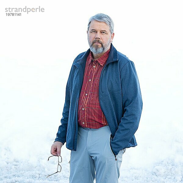 Mann  56 Jahre alt mit grauen Haaren und grauem Vollbart steht vor einer Schneefläche im Winter  rot kariertes Hemd  dunkelblaue Jacke  hellgraue Hose  Brille in der Hand  eine Hand in der Hosentasche  weißer Hintergrund