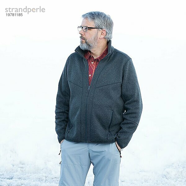 Mann  56 Jahre alt mit grauen Haaren und grauem Vollbart steht vor einer Schneefläche im Winter  graue Jacke  rotes Hemd  Hände in den Hosentaschen  Blick zur Seite  mit Brille  weißer Hintergrund