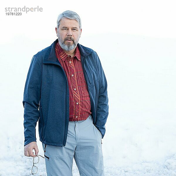 Mann  56 Jahre alt mit grauen Haaren und grauem Vollbart steht vor einer Schneefläche im Winter  rot kariertes Hemd  dunkelblaue Jacke  hellgraue Hose  Brille in der Hand  eine Hand in der Hosentasche  weißer Hintergrund