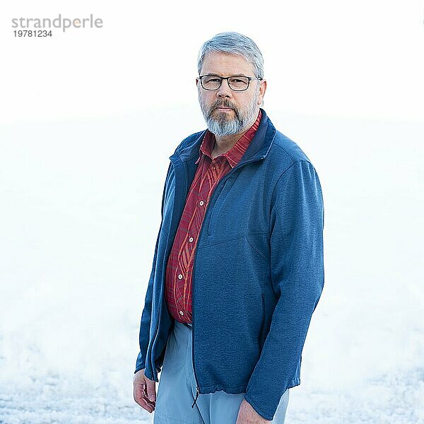 Mann  56 Jahre alt mit grauen Haaren und grauem Vollbart steht vor einer Schneefläche im Winter  rot kariertes Hemd  dunkelblaue Jacke  hellgraue Hose  mit Brille  weißer Hintergrund