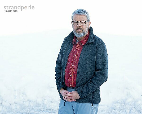 Mann  56 Jahre alt mit grauen Haaren und grauem Vollbart steht vor einer Schneefläche im Winter  Hände gefaltet  graue Jacke  rot kariertes Hemd  mit Brille  weißer Hintergrund