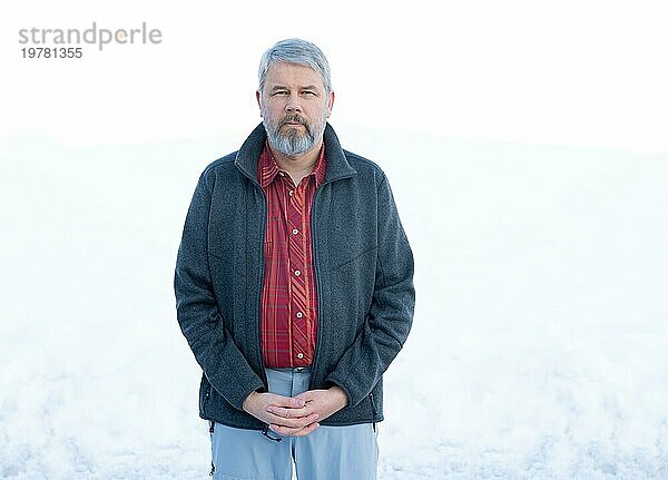 Mann  56 Jahre alt mit grauen Haaren und grauem Vollbart steht vor einer Schneefläche im Winter  Hände gefaltet  graue Jacke  rot kariertes Hemd  weißer Hintergrund