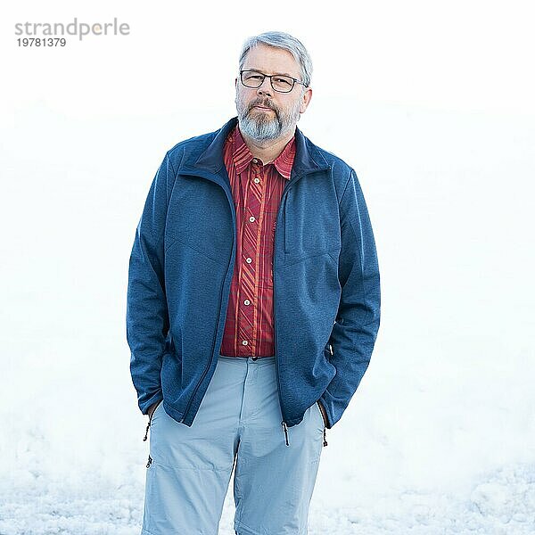 Mann  56 Jahre alt mit grauen Haaren und grauem Vollbart steht vor einer Schneefläche im Winter  rot kariertes Hemd  dunkelblaue Jacke  hellgraue Hose  Hände in den Hosentaschen  mit Brille  weißer Hintergrund