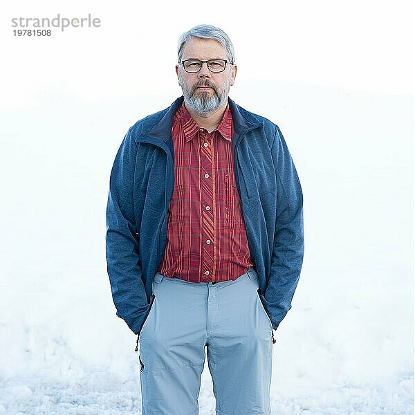 Mann  56 Jahre alt mit grauen Haaren und grauem Vollbart steht vor einer Schneefläche im Winter  rot kariertes Hemd  dunkelblaue Jacke  hellgraue Hose  Hände in den Hosentaschen  kritischer Blick  mit Brille  weißer Hintergrund