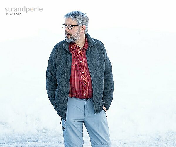 Mann  56 Jahre alt mit grauen Haaren und grauem Vollbart steht vor einer Schneefläche im Winter  Hände in den Hosentaschen  graue Jacke  rot kariertes Hemd  Blick zur Seite  mit Brille  weißer Hintergrund