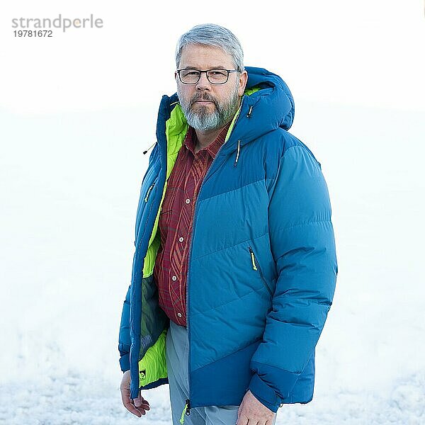 Mann  56 Jahre alt mit grauen Haaren und grauem Vollbart steht vor einer Schneefläche im Winter  dicke blaue Daunenjacke  rot kariertes Hemd  mit Brille  weißer Hintergrund