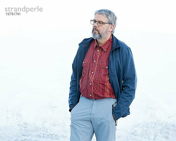 Mann  56 Jahre alt mit grauen Haaren und grauem Vollbart steht vor einer Schneefläche im Winter  rot kariertes Hemd  dunkelblaue Jacke  hellgraue Hose  Hände in den Hosentaschen  Blick zur Seite  mit Brille  weißer Hintergrund