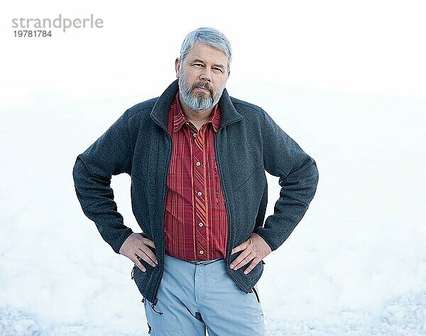 Mann  56 Jahre alt mit grauen Haaren und grauem Vollbart steht vor einer Schneefläche im Winter  Kopf zur Seite geneigt  Hände in die Hüften gestemmt  graue Jacke  rot kariertes Hemd  weißer Hintergrund