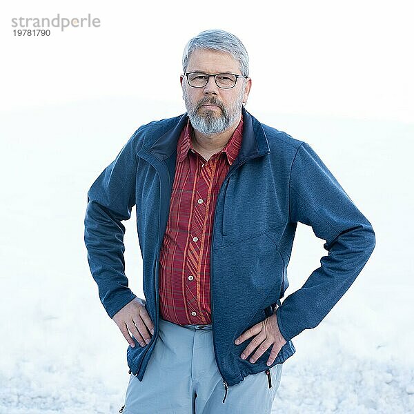 Mann  56 Jahre alt mit grauen Haaren und grauem Vollbart steht vor einer Schneefläche im Winter  rot kariertes Hemd  dunkelblaue Jacke  hellgraue Hose  Hände in die Seiten gestemmt  mit Brille  weißer Hintergrund