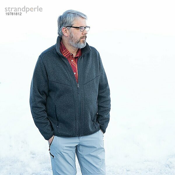 Mann  56 Jahre alt mit grauen Haaren und grauem Vollbart steht vor einer Schneefläche im Winter  graue Jacke  rotes Hemd  Hände in den Hosentaschen  Blick zur Seite  mit Brille