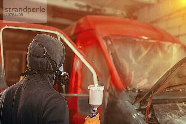 Karosseriereparatur eines Lieferwagens  Lackierung der Karosserie in Rot  ein Mechaniker mit Atemschutzmaske lackiert einen Lieferwagen mit einer Spritzpistole