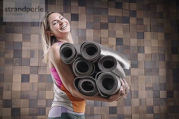 Bild einer fröhlichen erwachsenen Frau  die einen Stapel gerollter Übungsmatten trägt. Yoga und Pilates Konzept. Training  Tanzen und Posieren. Gemischte Medien