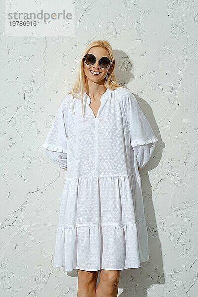 Zufriedene blonde Frau mit Sonnenbrille und weißem Sommerkleid an die Wand gedrückt