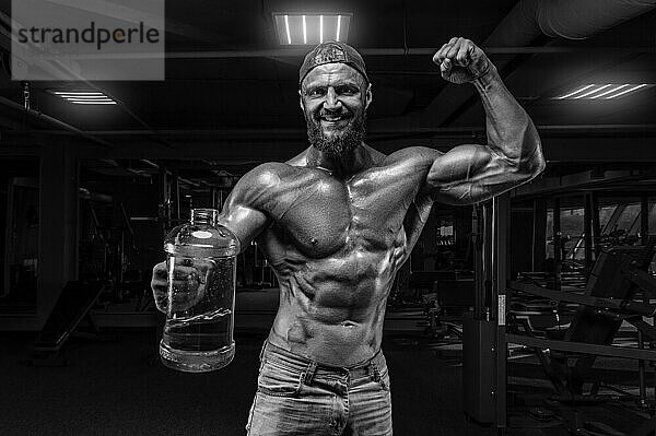 Muskulöser Mann steht in der Turnhalle mit einer großen Flasche Sportnahrung. Fitness und Bodybuilding Konzept. Gemischte Medien