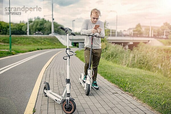 Junger gutaussehender Blondschopf  der sich die Route auf seinem Smartphone ansieht  um mit einem Freund einen Ausflug mit dem Elektroroller zu machen