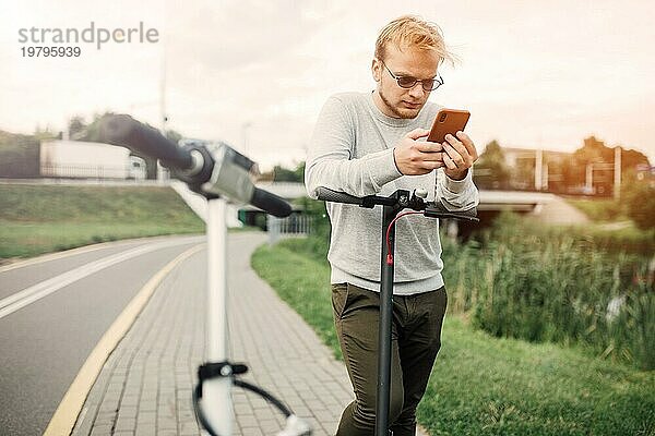 Junger gutaussehender Blondschopf  der sich die Route auf seinem Smartphone ansieht  um mit einem Freund einen Ausflug mit dem Elektroroller zu machen