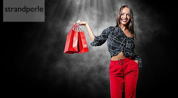 Schöne glückliche Brünette mit roten Paketen in ihren Händen. Schwarzer Hintergrund. Konzept für Rabatte  Verkäufe  Einkäufe. Gemischte Medien