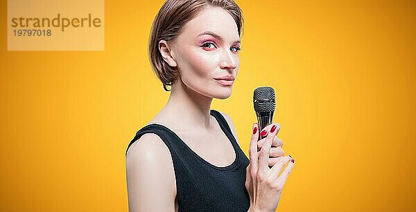 Porträt einer eleganten  stilvollen Frau mit einem Mikrofon. Gelber Hintergrund. Karaoke Konzept.