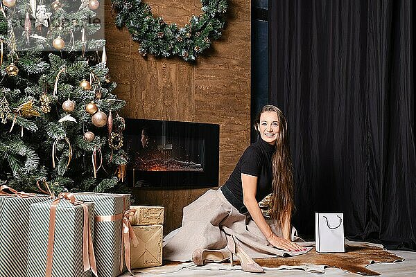 Eine fröhliche Frau sitzt auf dem Boden neben einem warmen Kamin  der mit einem üppigen grünen Kranz geschmückt ist. Die Atmosphäre ist gemütlich und intim und fängt das Wesen der Weihnachtswärme und Freude ein