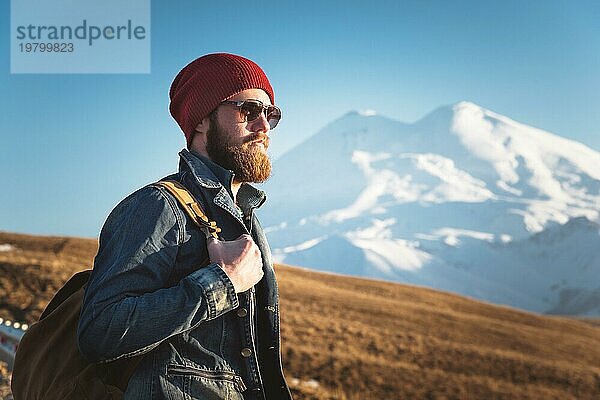 Porträt eines nachdenklichen stilvollen Hipsters mit Bart  Sonnenbrille und Hut mit einem Rucksack vor dem Hintergrund des schneebedeckten Berges Elbrus im Kaukasus bei Sonnenuntergang