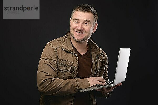Fröhlicher verrückter kaukasischer Mann  der lächelt und Grimassen schneidet  benutzt einen Laptop. Studioaufnahme vor schwarzem Hintergrund. Benutzer von Chats und Korrespondenz