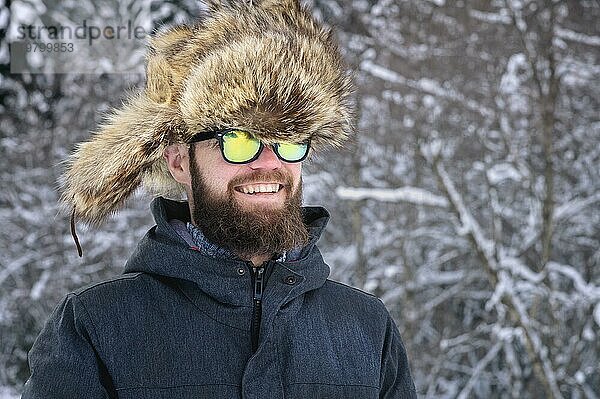 Porträt eines attraktiven jungen bärtigen Mannes mit einer großen Pelzmütze und einer Sonnenbrille Lacht über den verschneiten Wald in den Bergen