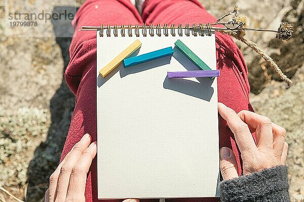 Blick aus der ersten Person auf den weiblichen Schoß ist ein Notizbuch mit Kreiden für die Zeichnung Pastelle. Weibliche Hände halten ein Notizbuch in der Natur