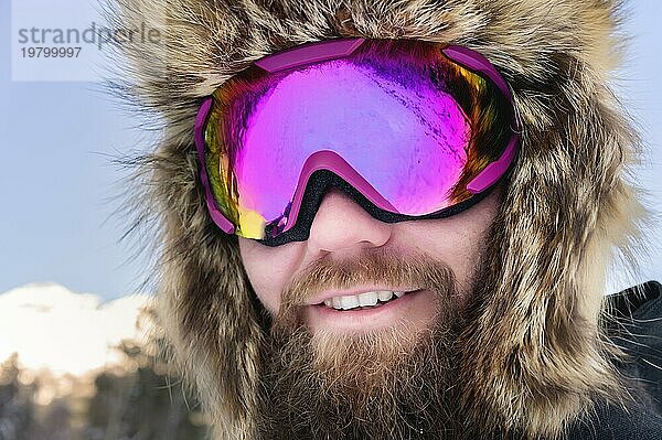 Close up Porträt eines bärtigen glücklichen Snowboarder Skifahrer in einer Skimaske mit Brille und einem Pelz großen Old School Hut. auf einem Hintergrund von einem Winter verschneiten Bergen