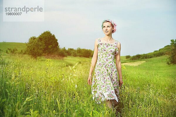 Junges glückliches lächelndes Mädchen in einem Kattunkleid mit einem Blumenstrauß geht eine Landstraße mit grünem Gras entlang