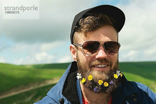 Nahaufnahme eines fröhlichen bärtigen Mannes mit Sonnenbrille und einer grauen Mütze mit Wildblumen im Bart. Sanfte Brutalität und gute Männlichkeit