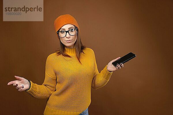 Eine schöne junge kaukasische Frau mit wallendem Haar in einem Pullover und einem Hut mit Brille hält ihr Smartphone in der Hand  breitet ihre Arme aus und blickt in die Kamera. verwirrt Studioporträt auf braunem Hintergrund. copy space